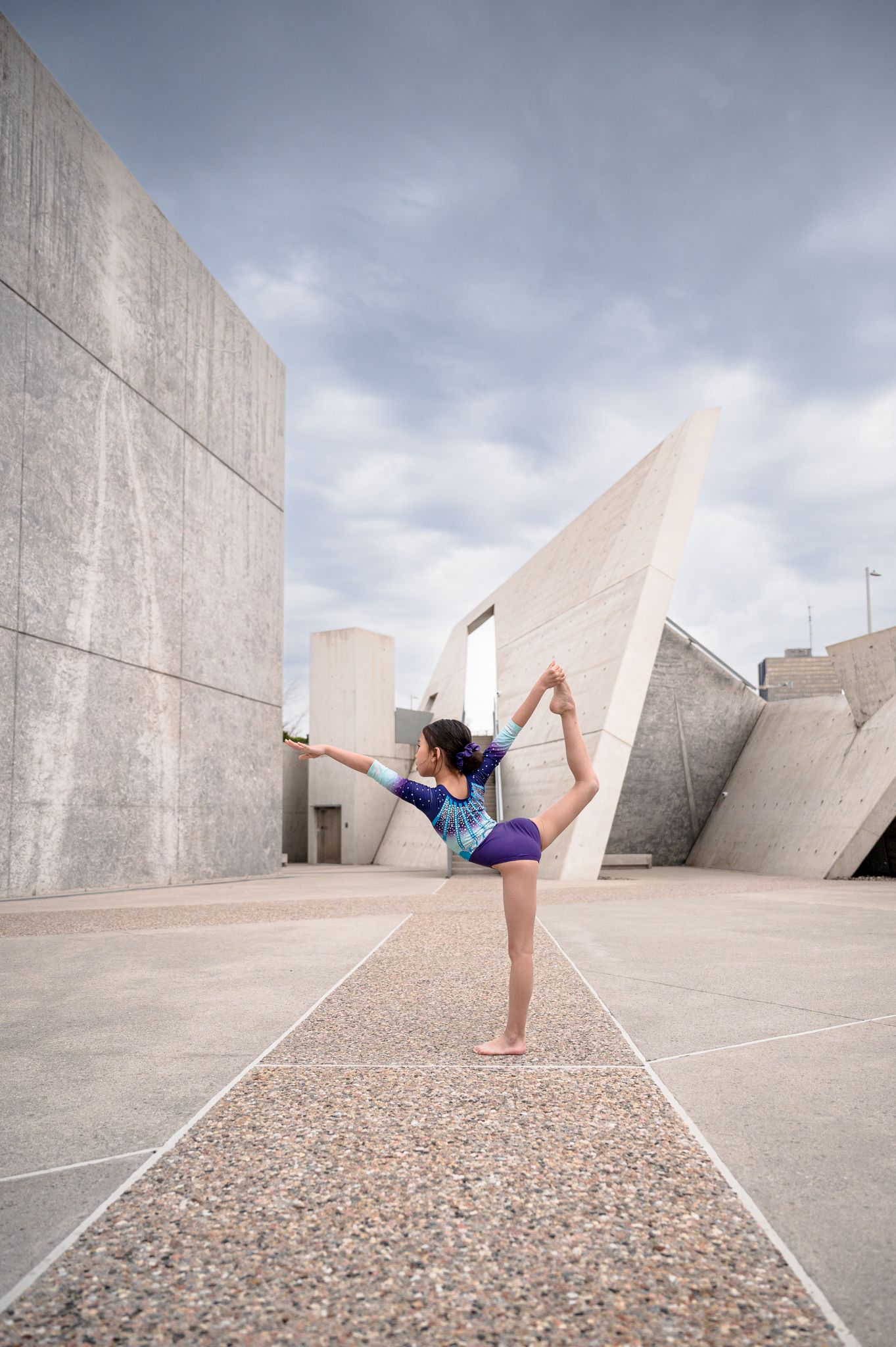 Dancer performing a graceful high kick pose against geometric concrete architecture under cloudy skies.