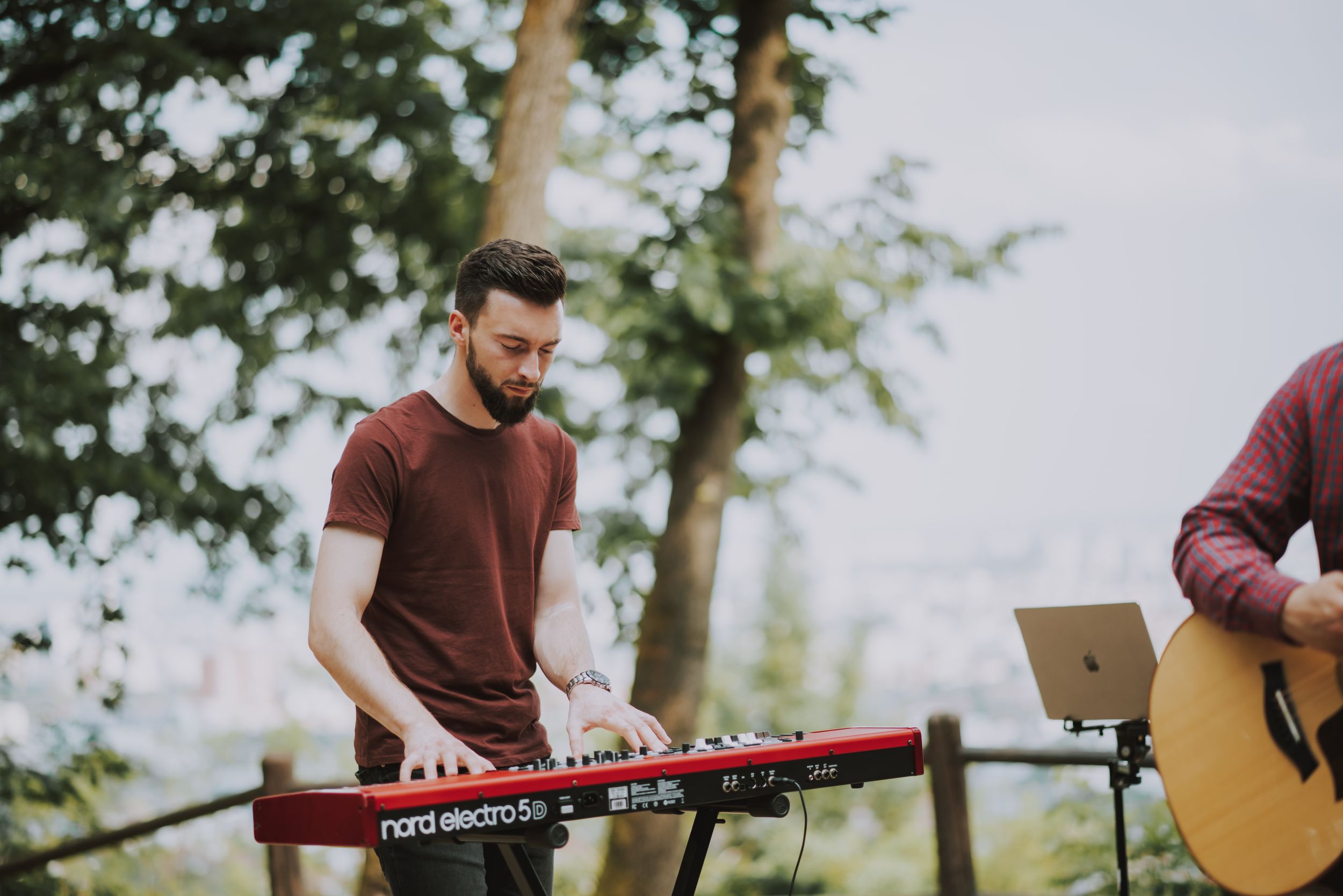 A keyboard player performs on a red Nord keyboard at an outdoor venue with trees in the background.