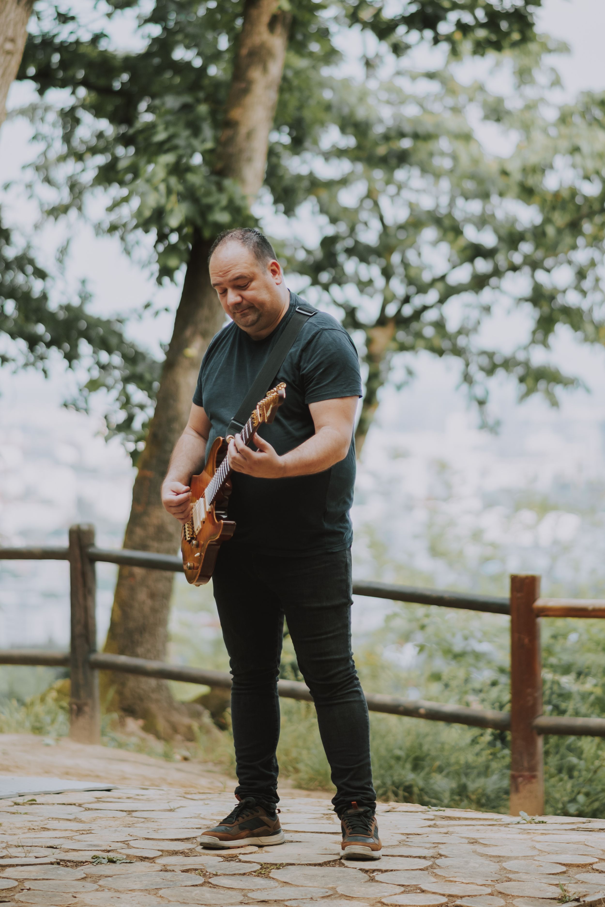 A musician playing an electric guitar performs outdoors on a scenic overlook with trees in the background.