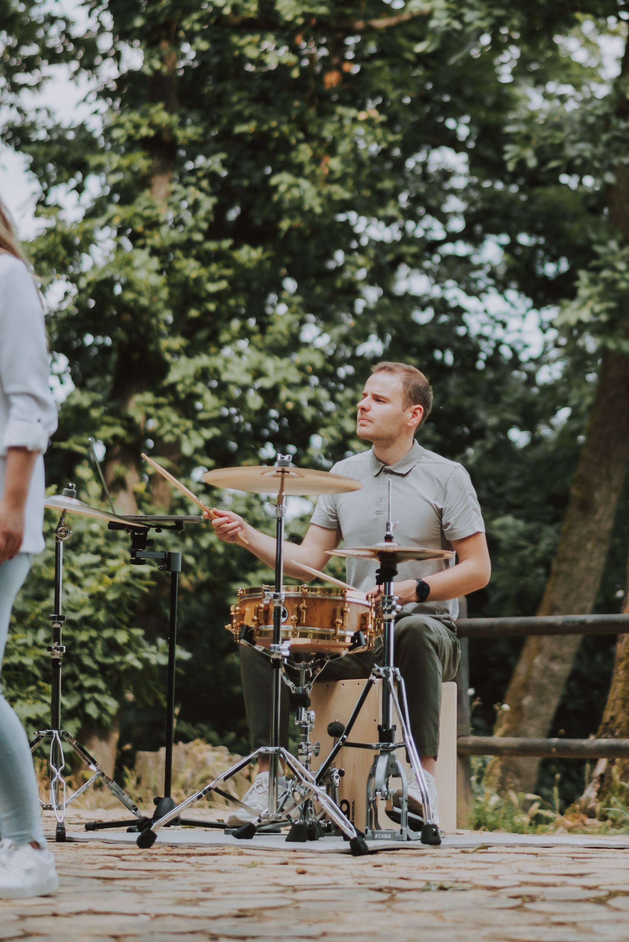 A drummer plays on a drum kit during an outdoor performance with leafy trees behind.