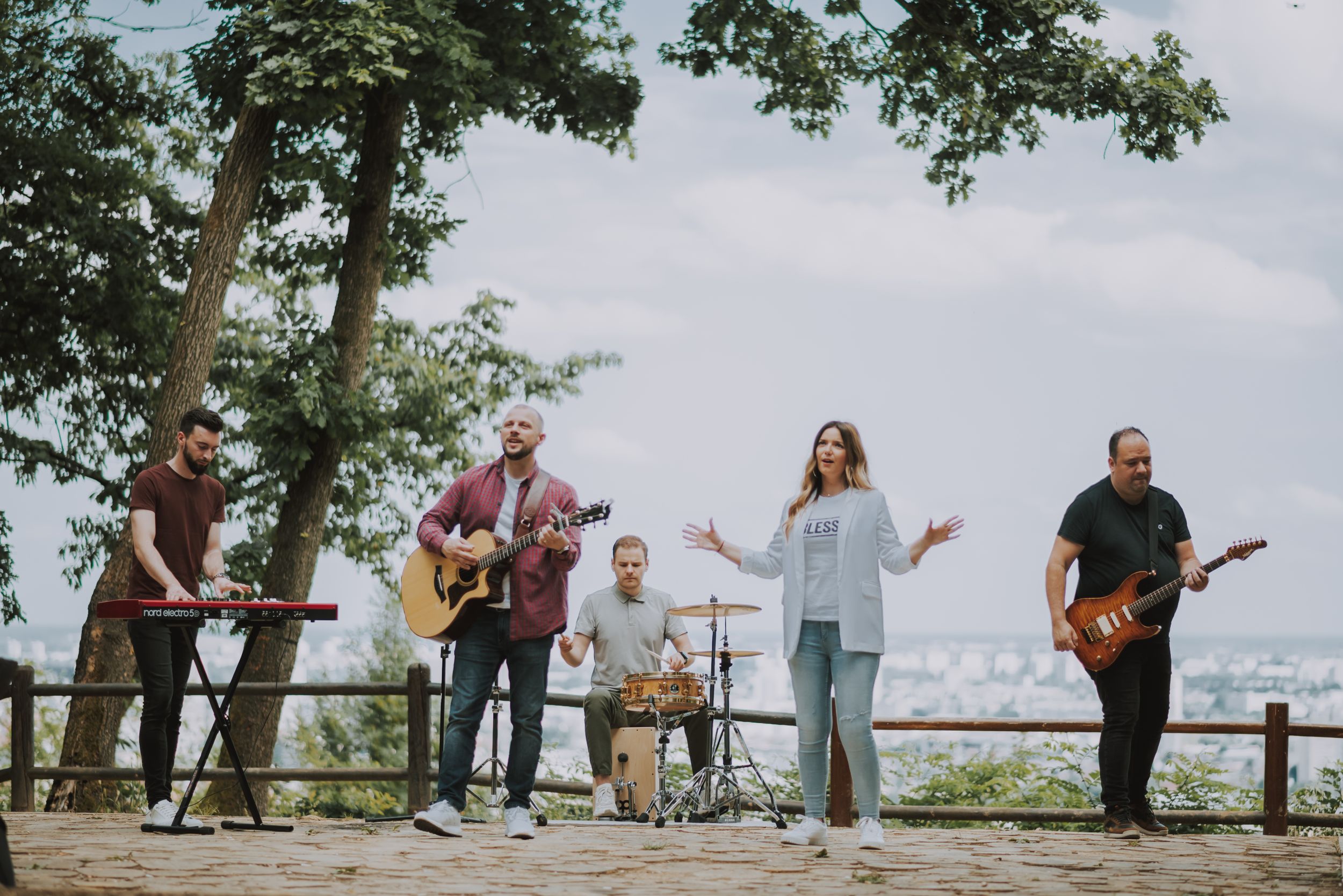 A band performs music together on a scenic overlook platform surrounded by trees with city views.