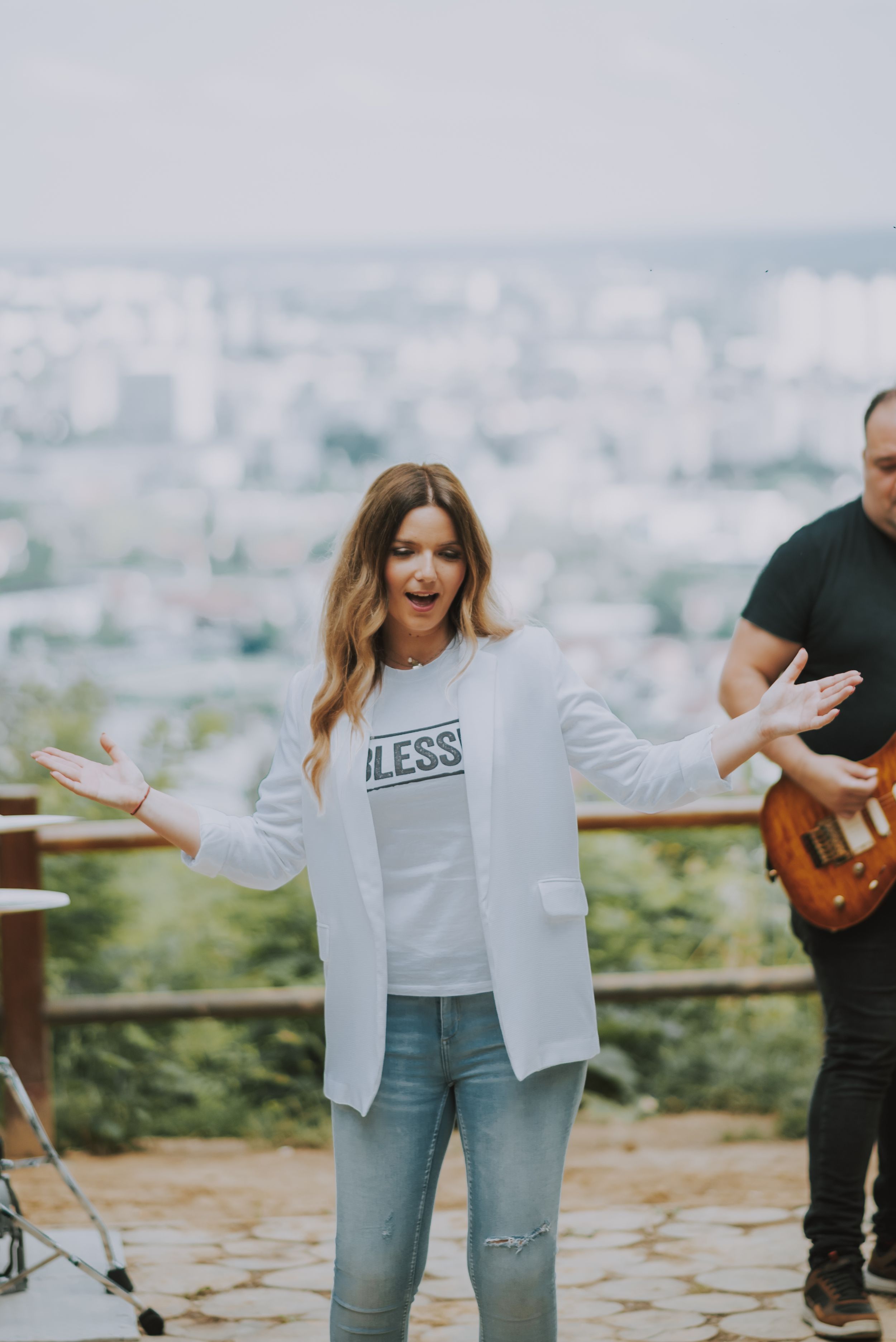 A musical performance on an outdoor overlook features singers and musicians with a cityscape backdrop.