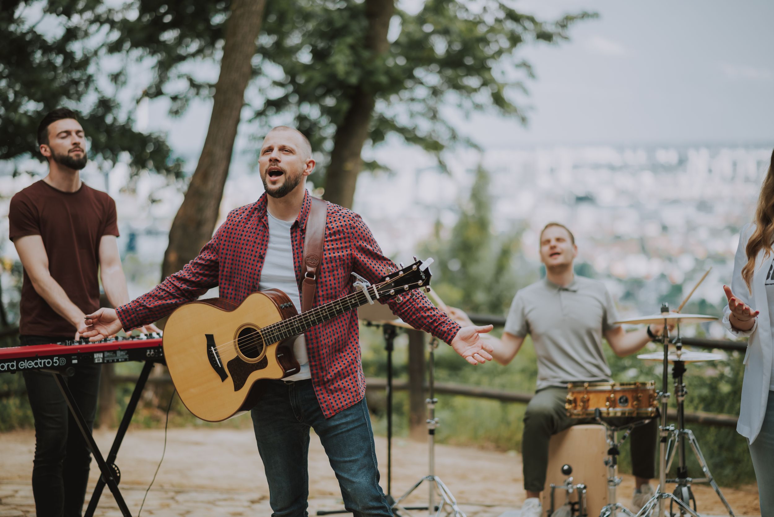 A worship band performs outdoors with acoustic guitar, keyboard and drums against a scenic overlook.