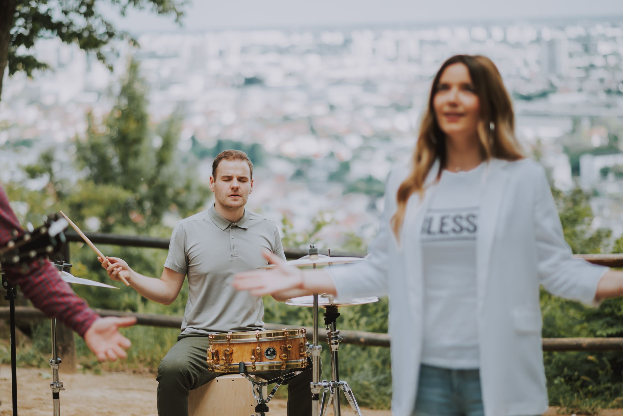Musicians perform on an elevated outdoor stage with drums, guitar and vocals against a city backdrop.