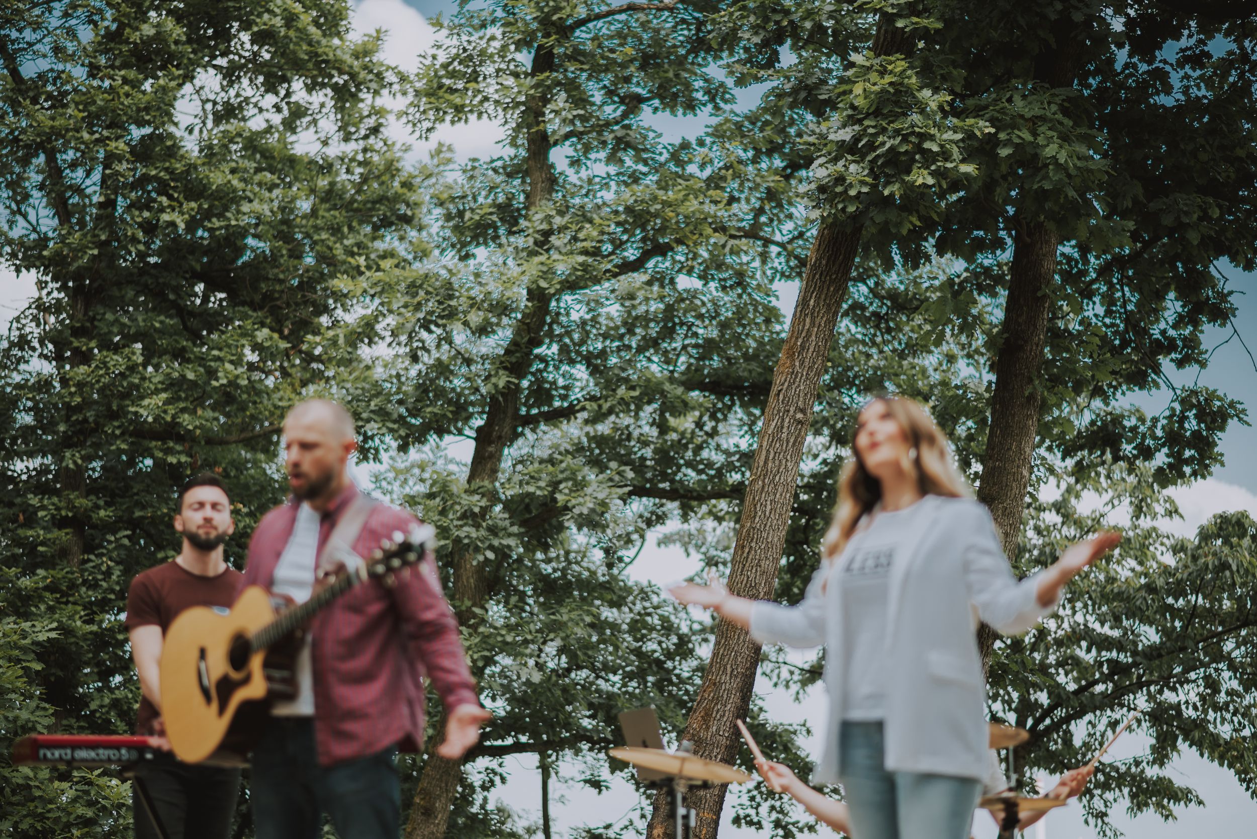 A music group plays instruments and sings during an outdoor performance surrounded by green trees.