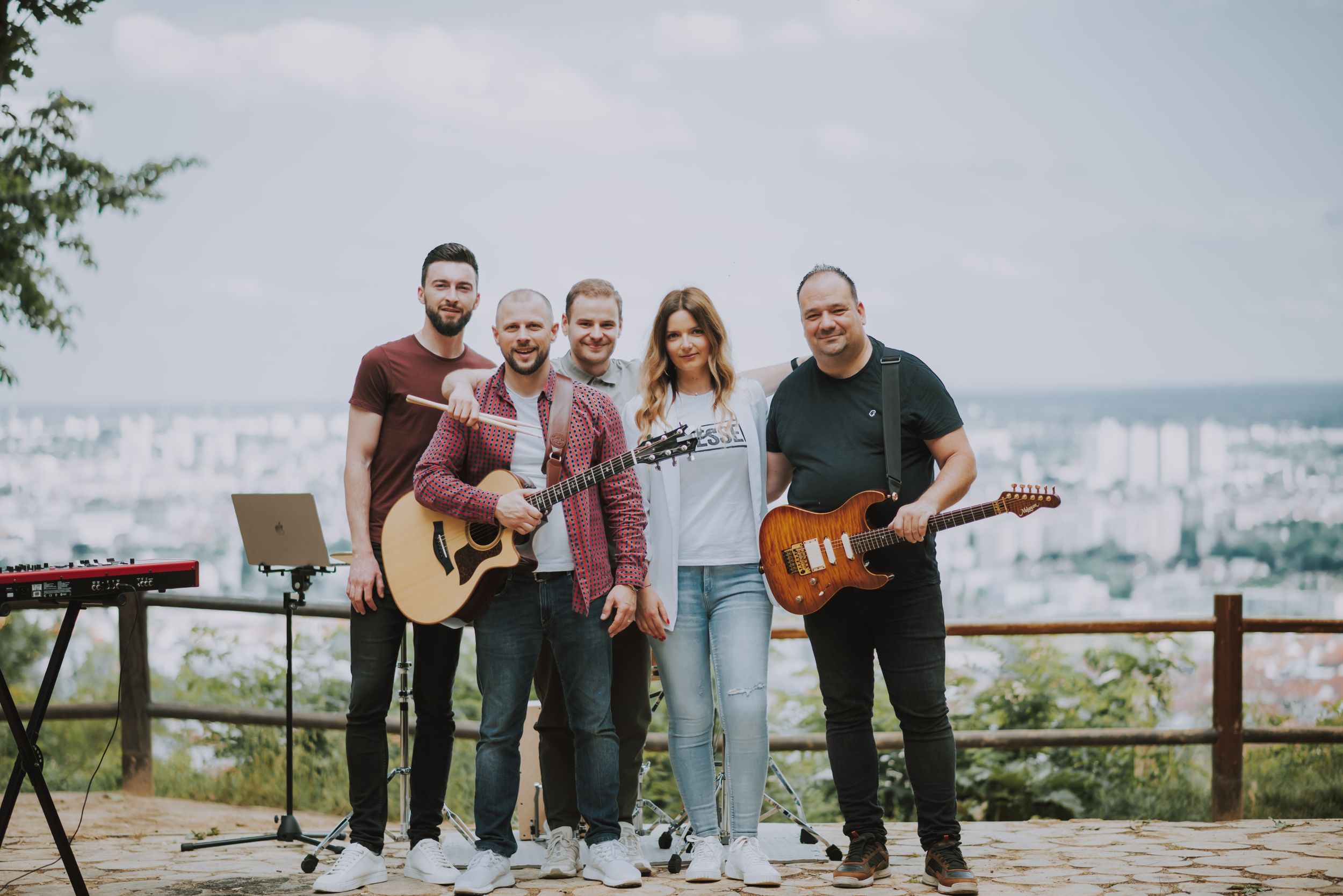 A five-piece band poses together for a group photo on an outdoor platform with city views behind them.