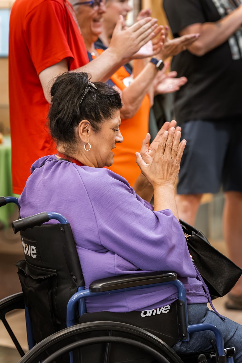 Woman in purple shirt clapping from wheelchair at indoor event, surrounded by standing applauding crowd.