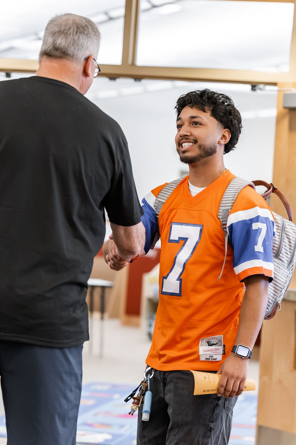 Young man in orange #7 jersey shaking hands with older man while wearing a backpack indoors.