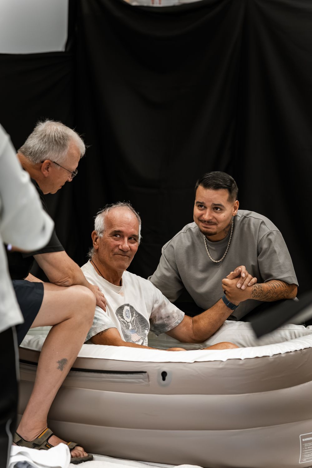 Three men gathered around an inflatable tub indoors, two sitting inside while a third leans in to talk.