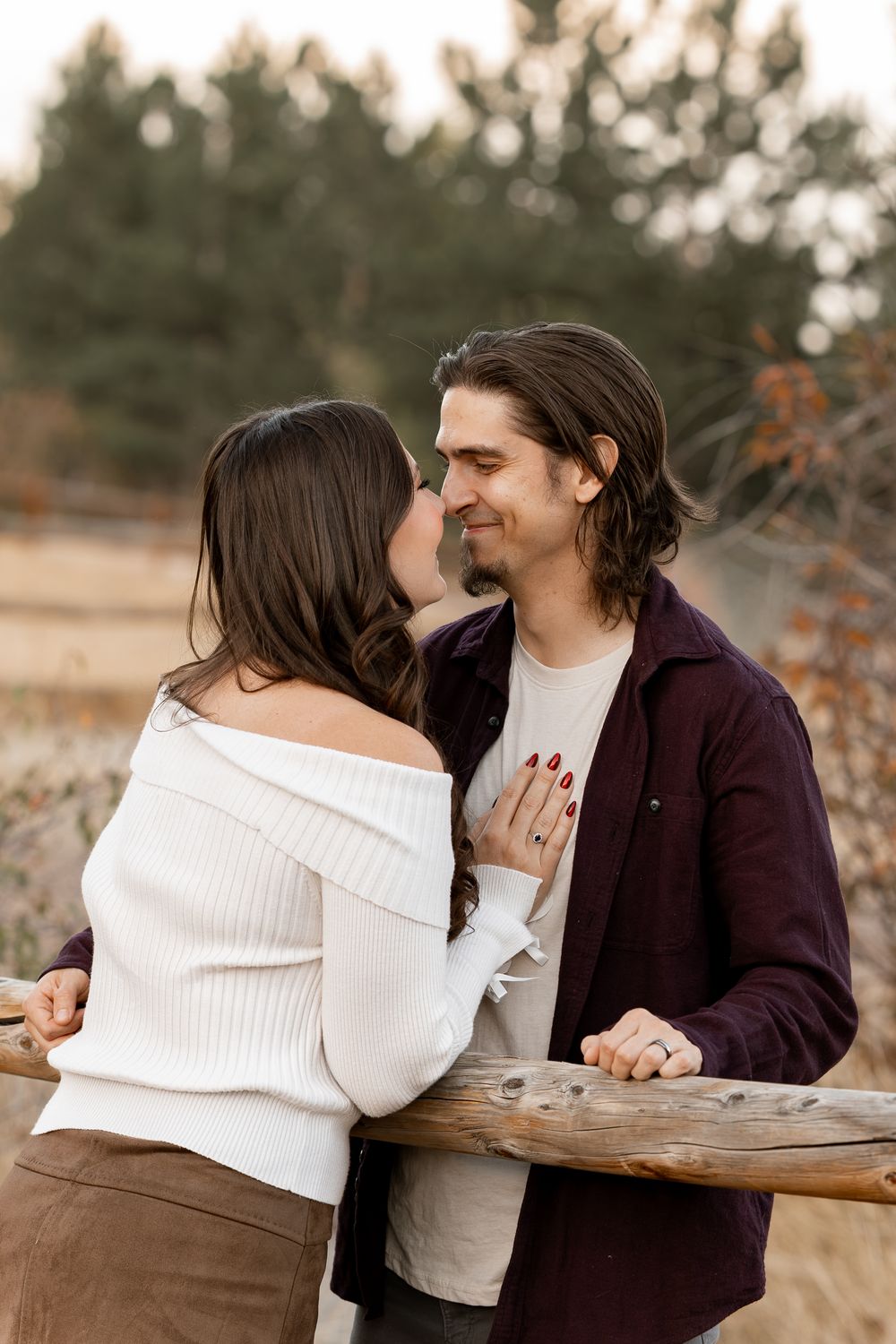 Couple sharing a romantic moment outdoors, woman in white off-shoulder sweater and man in burgundy shirt leaning on a wooden fence.