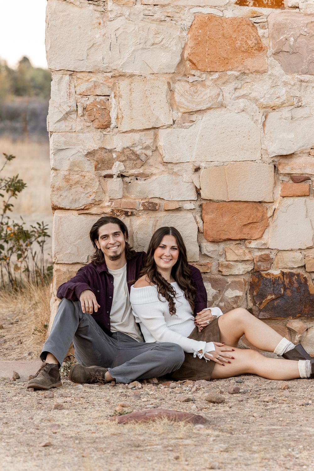 A smiling couple sits together against a rustic stone wall outdoors, dressed in casual fall attire.