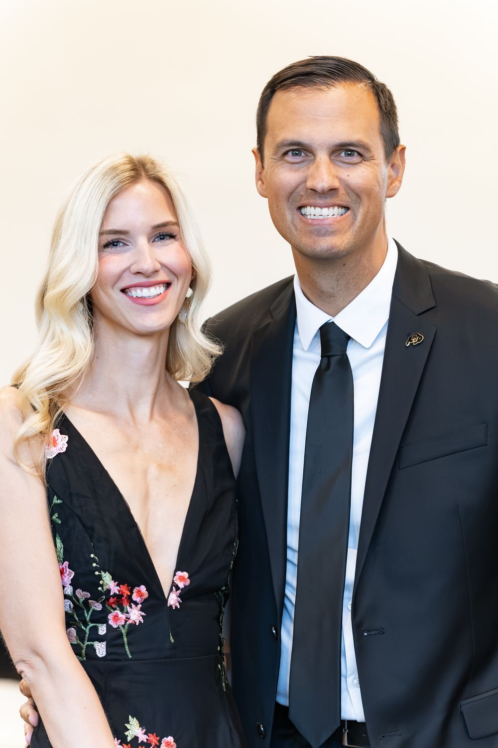 A smiling blonde woman in a black floral-embroidered dress poses beside a smiling man in a dark suit, white shirt and black tie.