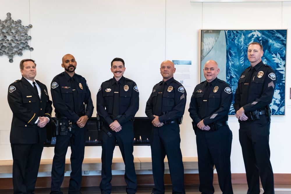 Six uniformed police officers pose side by side in a bright room with abstract wall art and a metal sculpture behind them.