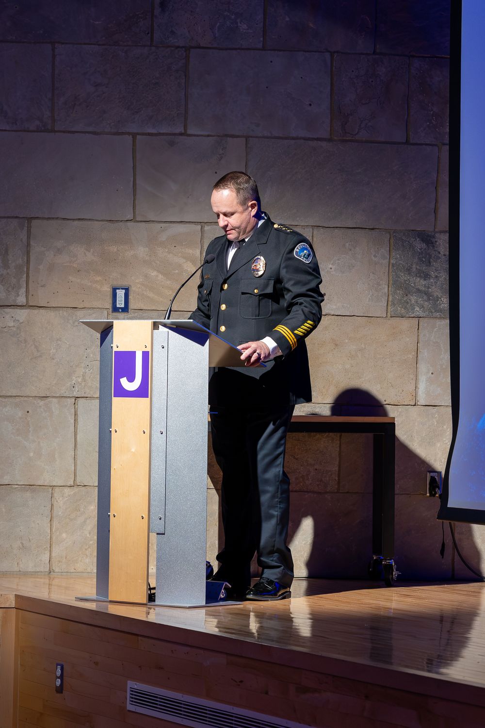 Police officer in dark uniform with gold stripes speaks at a wooden podium marked with a purple letter J on a stage before a stone wall.