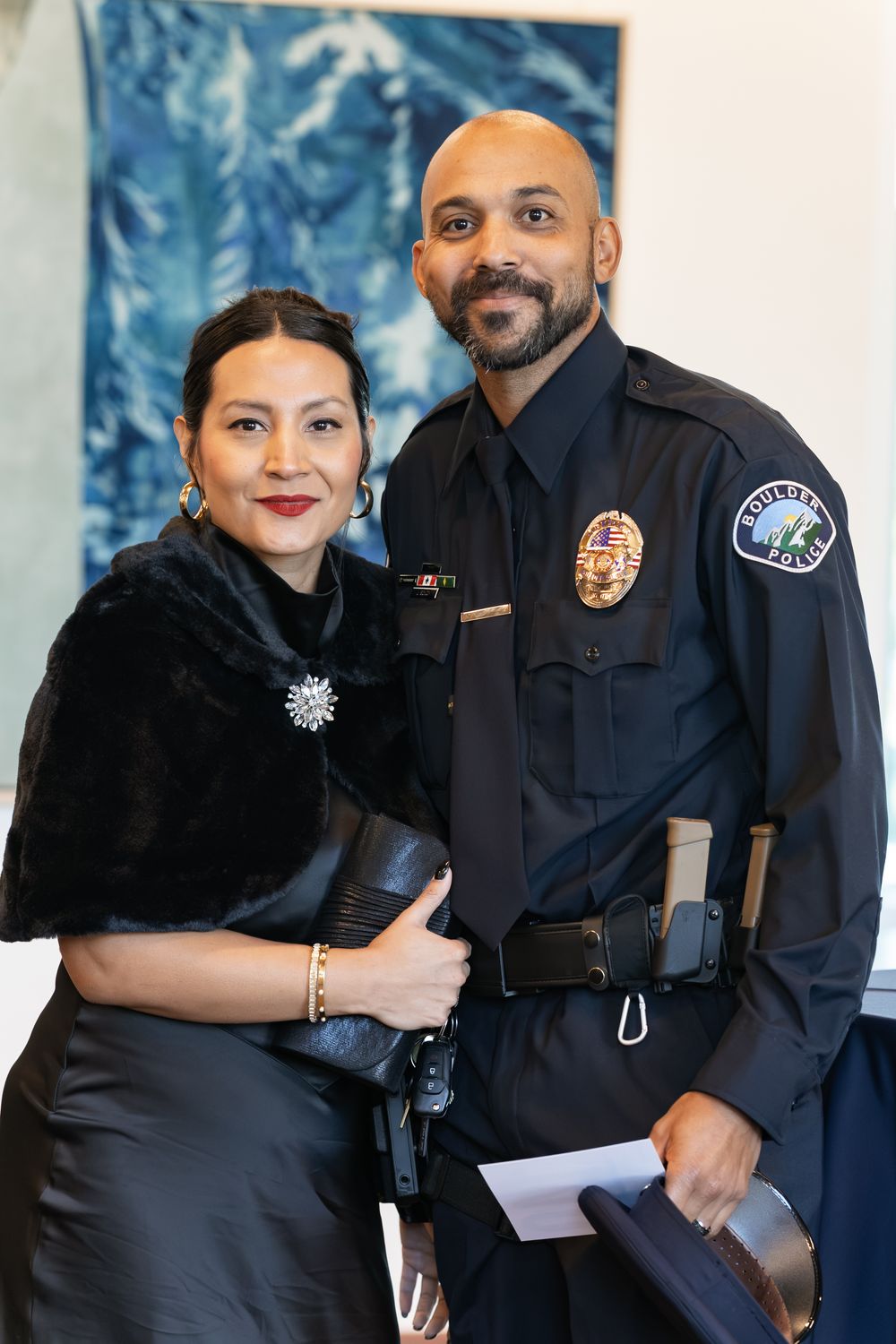 A smiling police officer in a dark uniform stands beside a woman in an elegant black outfit with a brooch at an indoor event.
