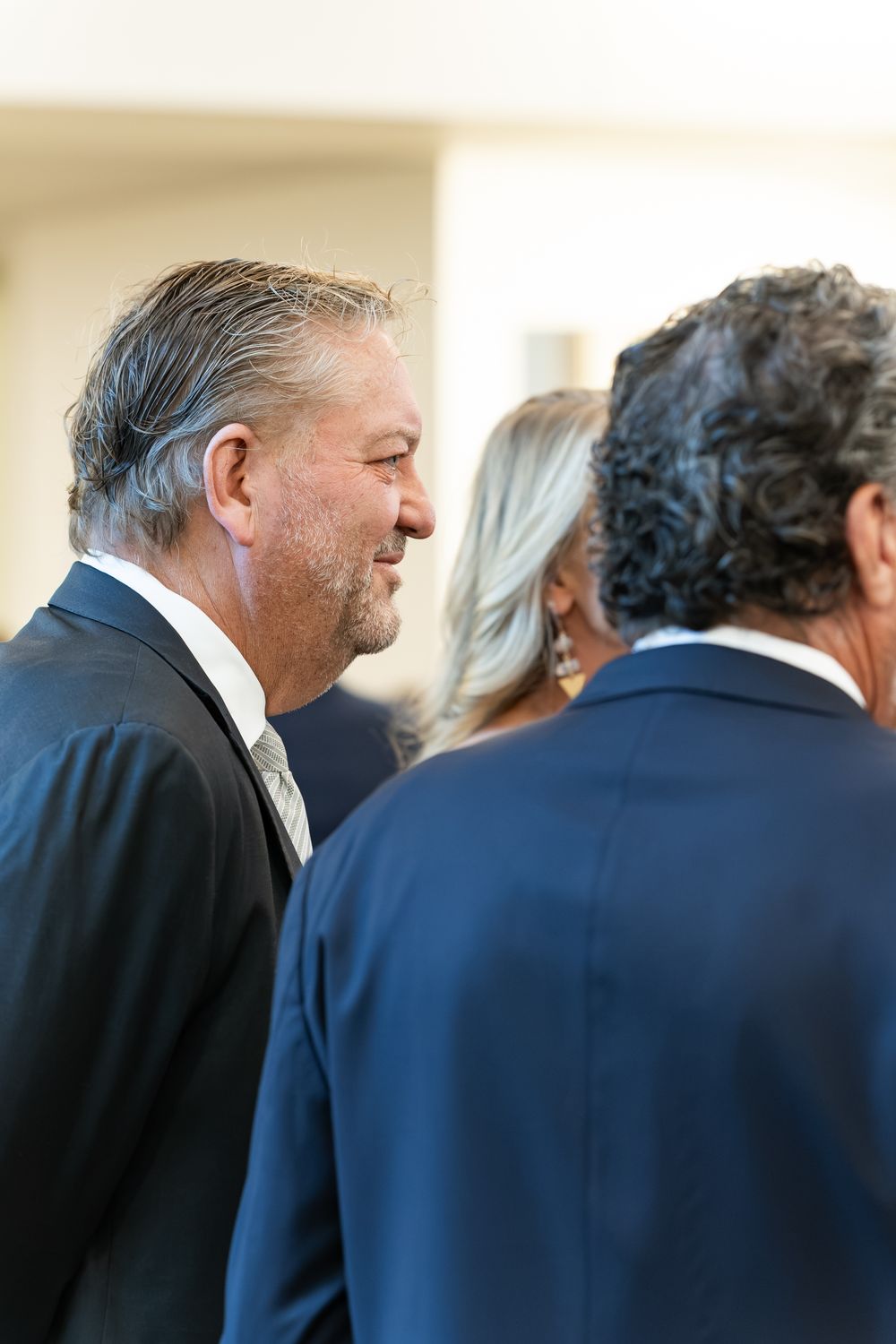 Smiling gray-haired man in a dark suit stands in profile at a social gathering, with a blonde woman and a man in a navy blazer nearby.