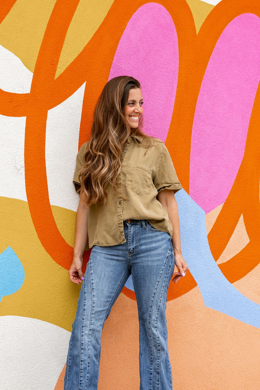 Woman in olive shirt and flare jeans smiling against colorful abstract mural wall.