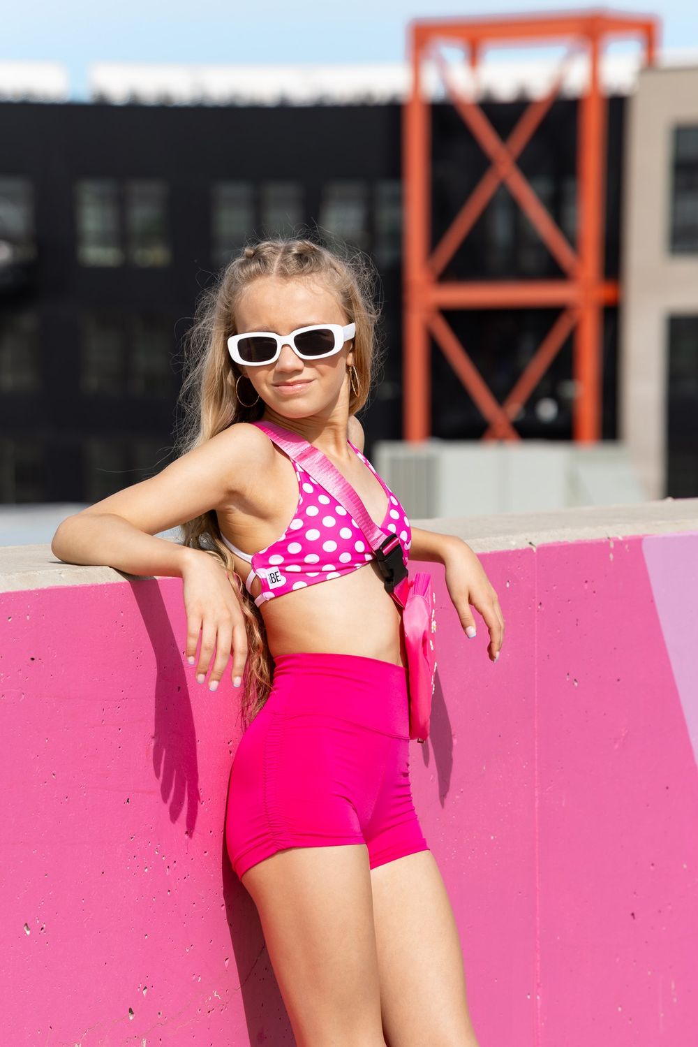 Young girl in pink polka dot bikini top and shorts with white sunglasses posing outdoors against a pink wall.