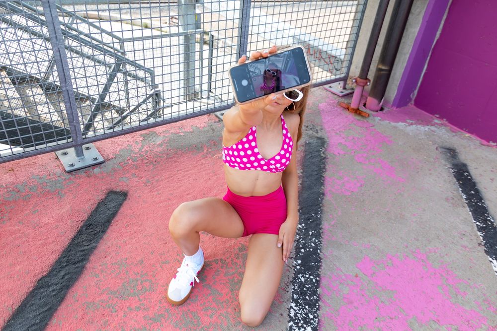 Woman in pink polka dot bikini taking a selfie with smartphone while kneeling on pink painted ground.