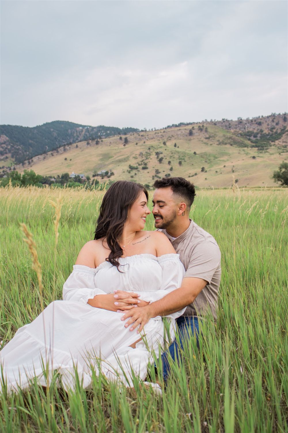 Couple in white and beige outfits sharing tender moments in tall grass with mountains behind them.