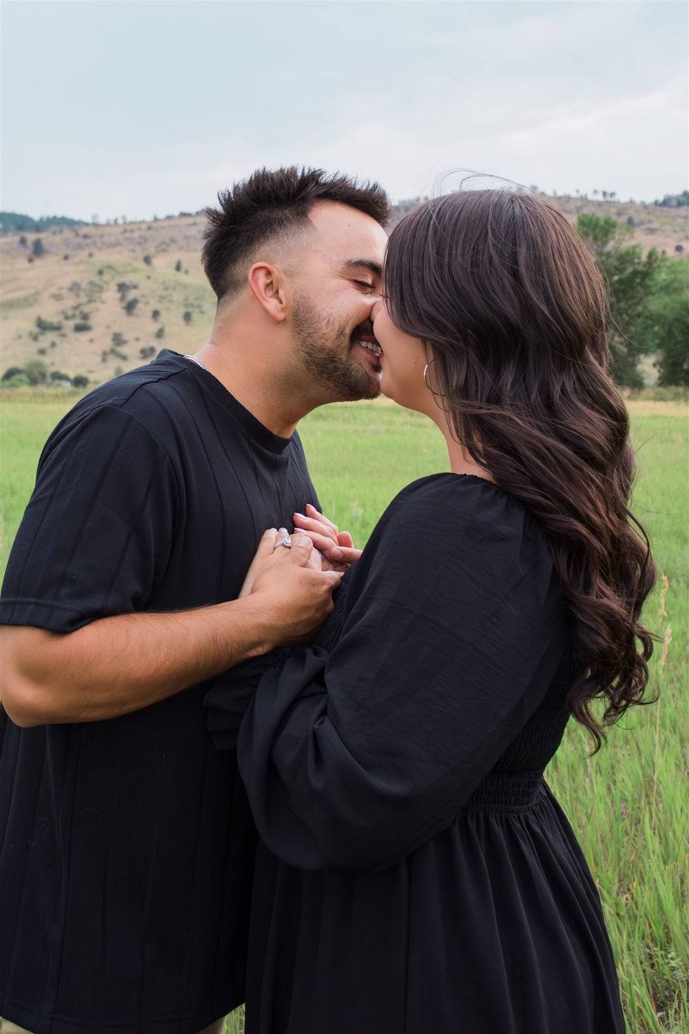 Romantic couple in black clothing embracing in a grassy field with mountains in the distance.
