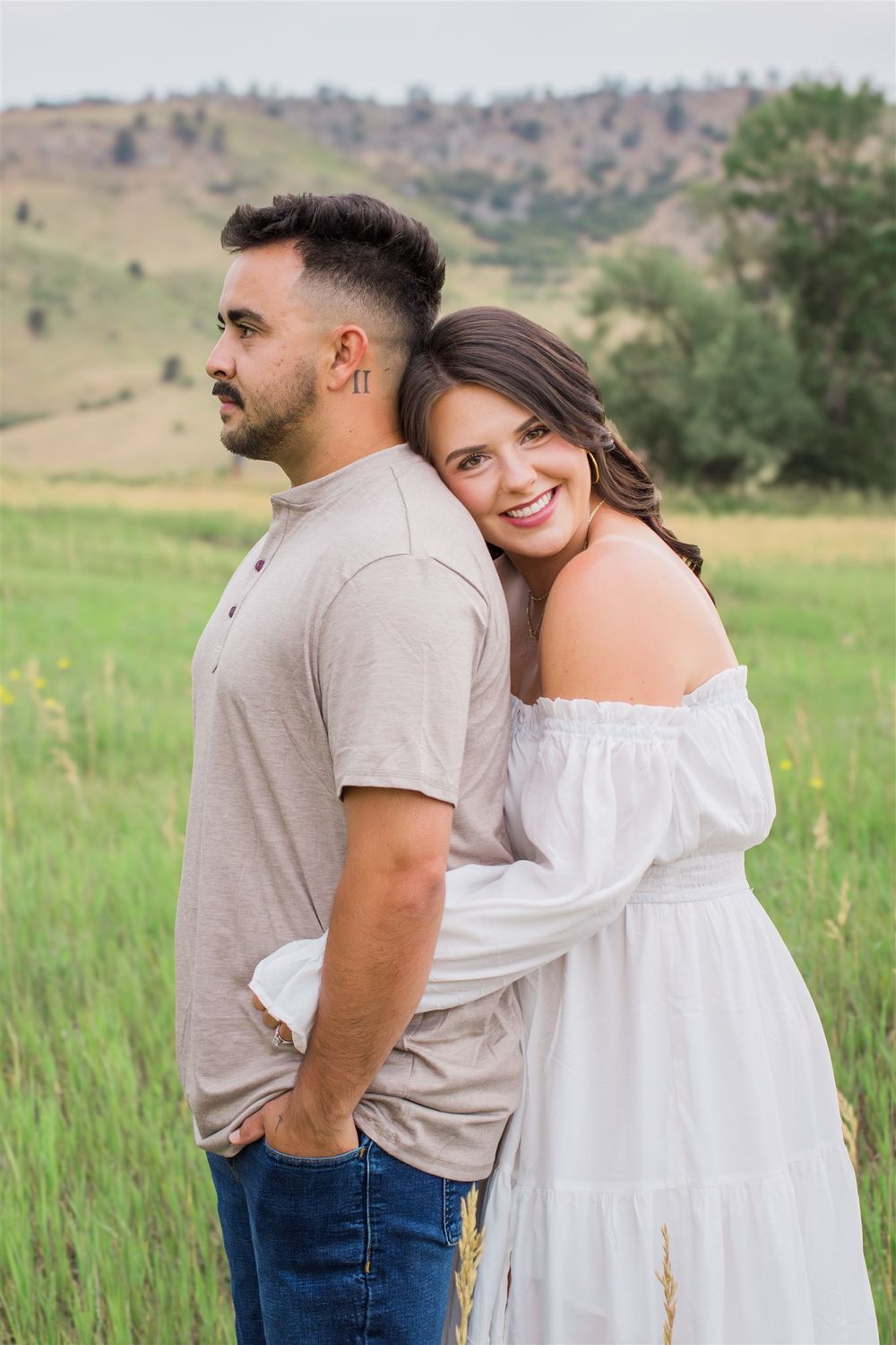 Couple poses in meadow wearing casual outfits with rolling hills in background.