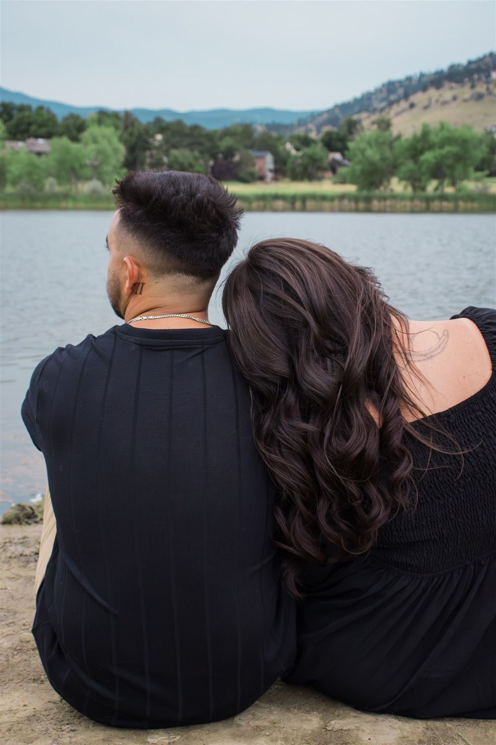 Couple in black clothing sitting together by a peaceful lake with mountain views.