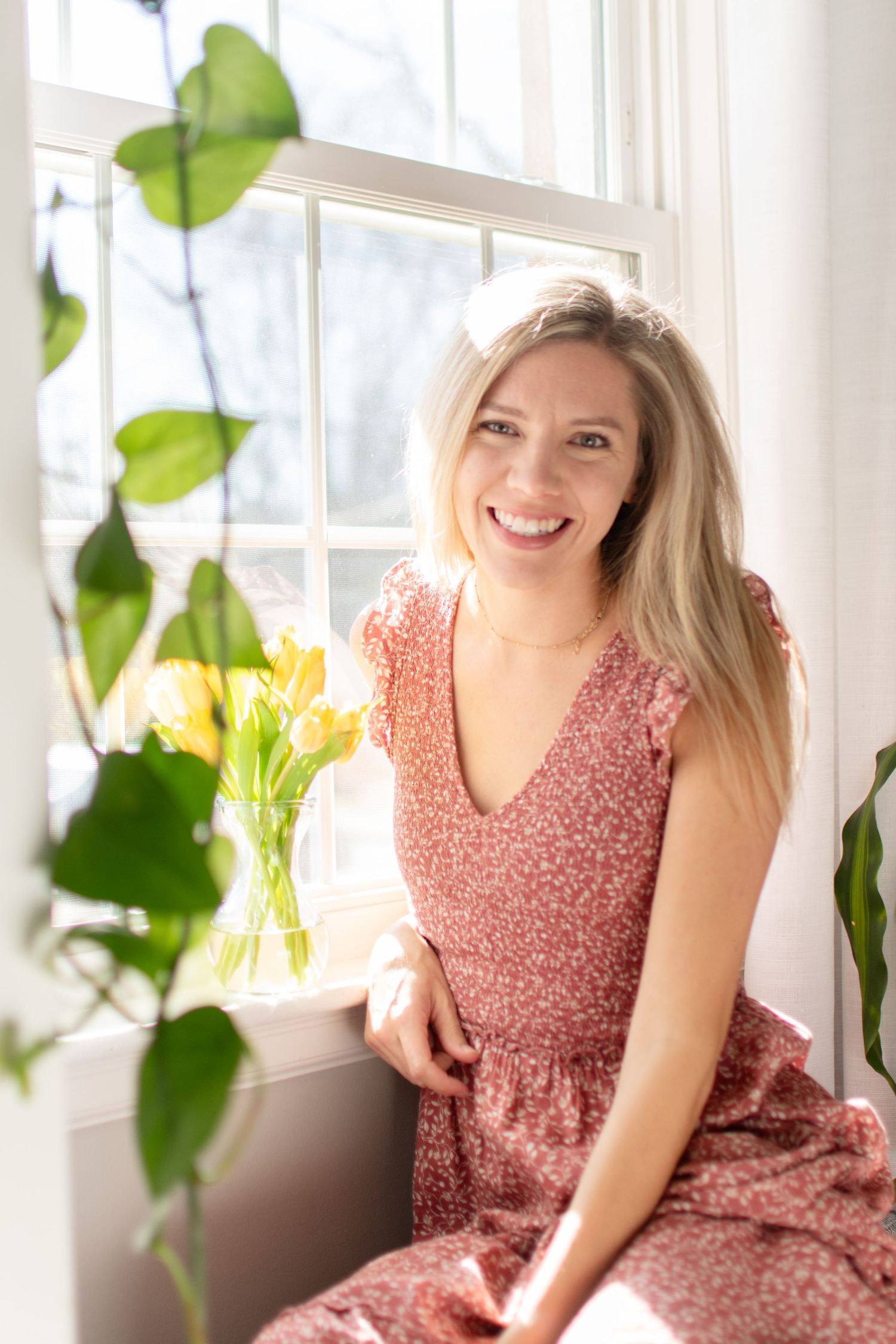 Pink floral dress against bright window with green plants and yellow tulips creating cheerful spring atmosphere.