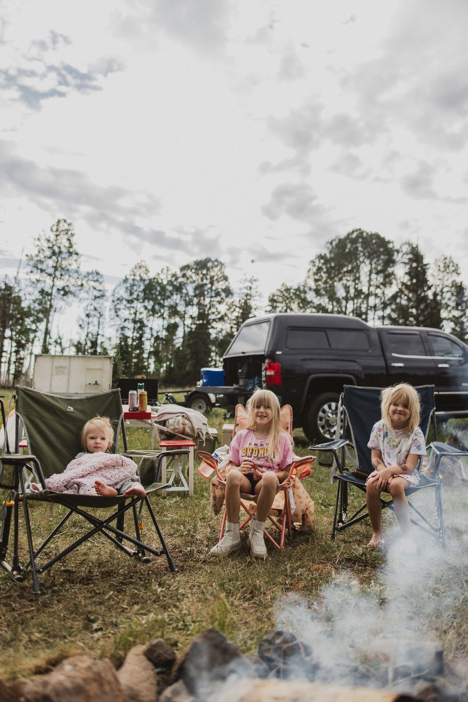 Campfire smoke rises near parked vehicles and camping chairs in forest setting.