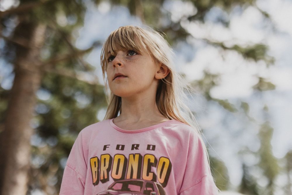 Child wearing a pink Bronco t-shirt stands against a forested background.