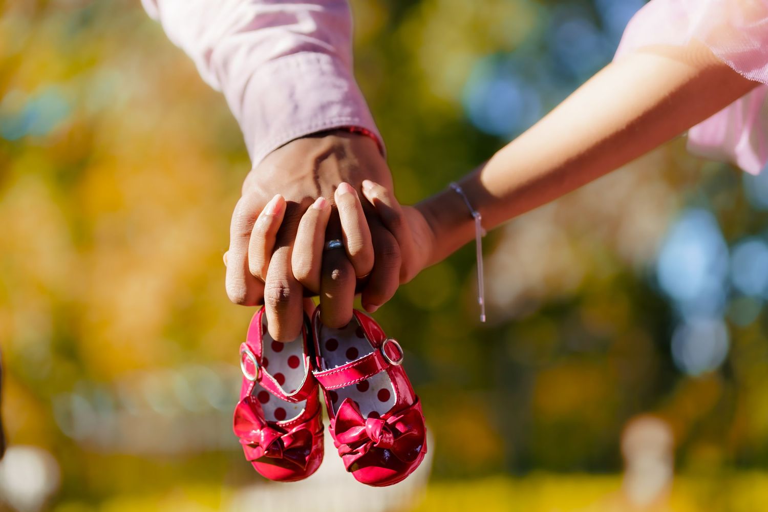 Close-up of hands holding tiny red polka dot baby shoes against blurred autumn background.