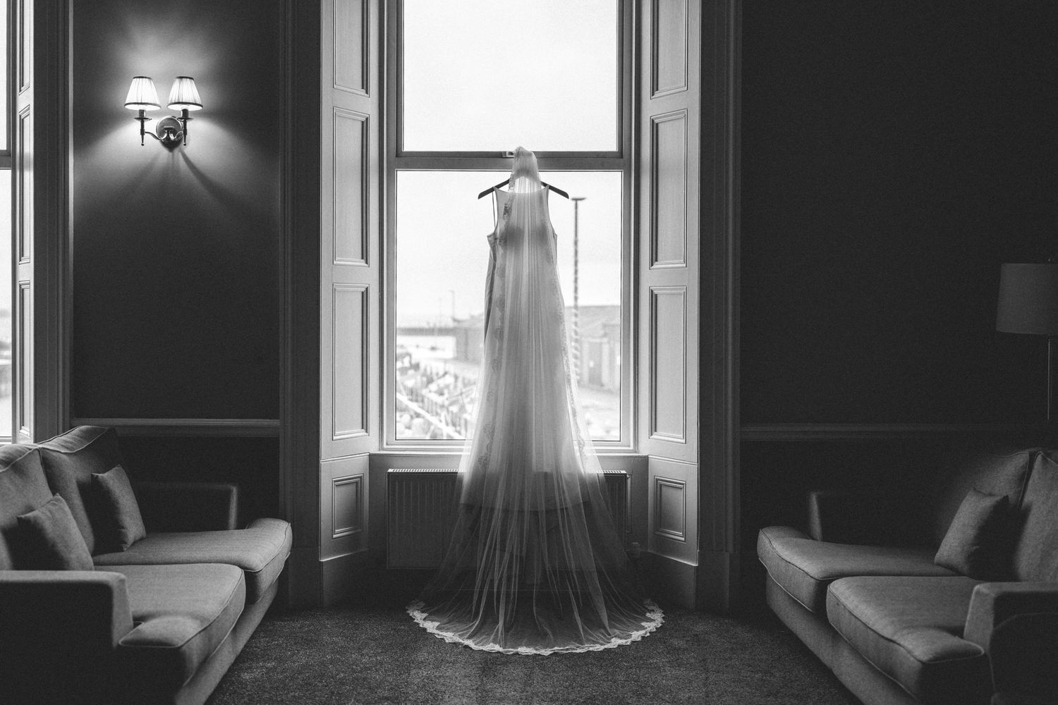 A dramatic black and white photograph of a wedding dress hanging in a doorway between two couches, backlit by natural light.