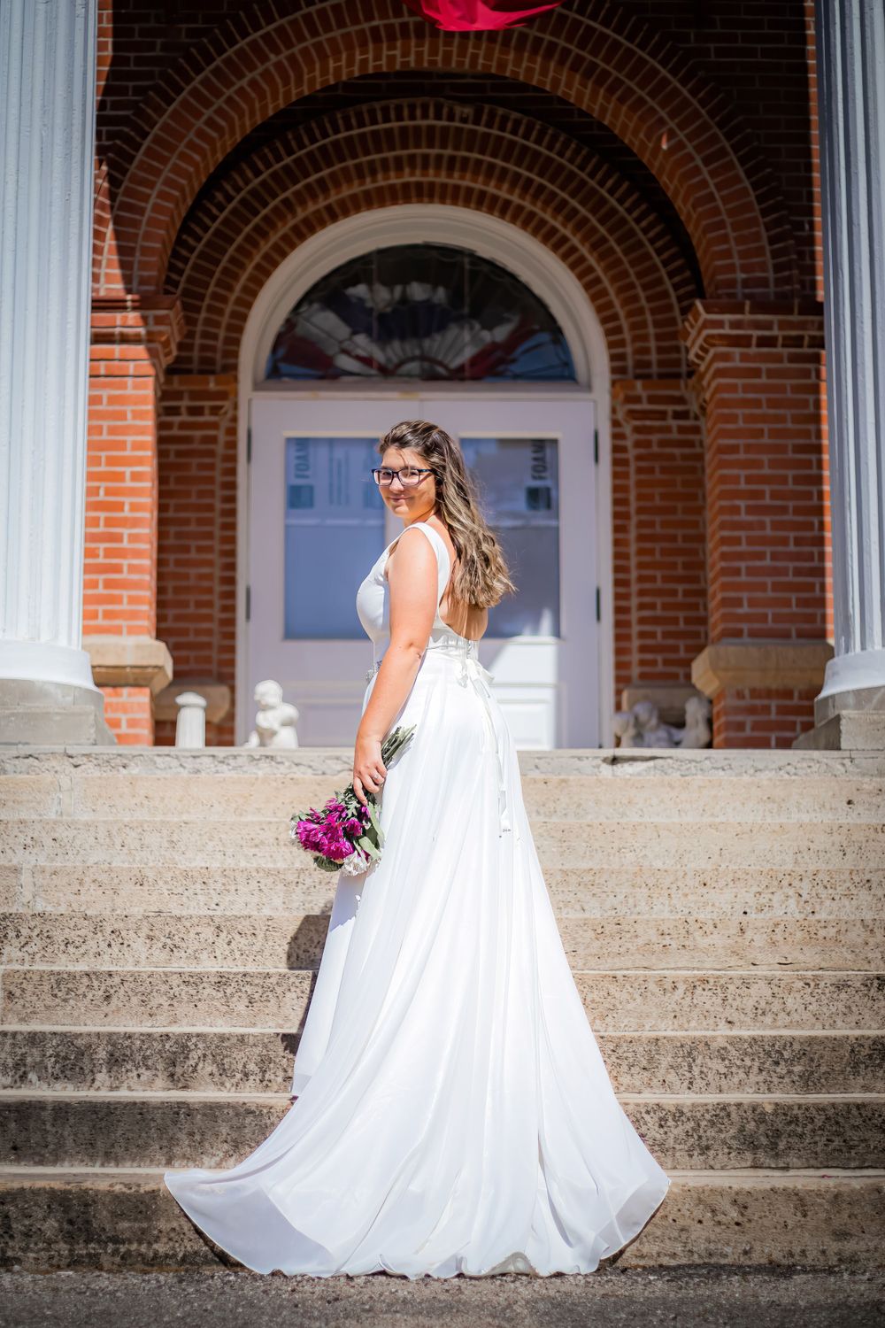 A bride in a flowing white gown poses on stone steps in front of a classic brick archway entrance.