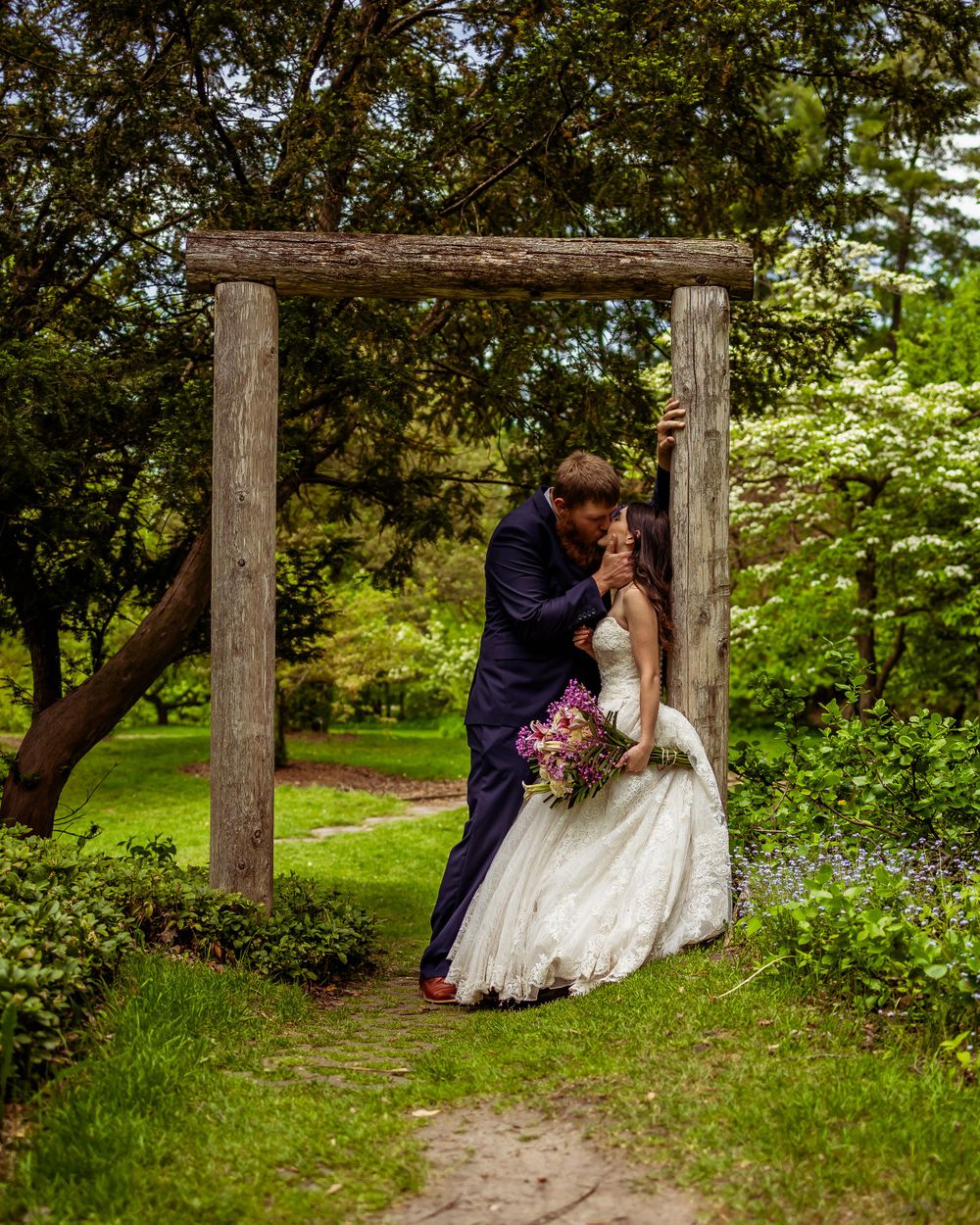 Romantic wedding couple shares kiss under rustic wooden arch in garden setting.