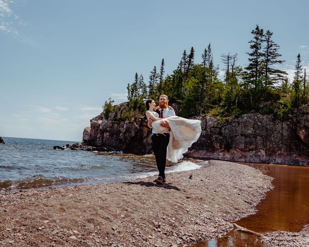 Groom carries bride playfully on beach with dramatic cliff coastline behind them.