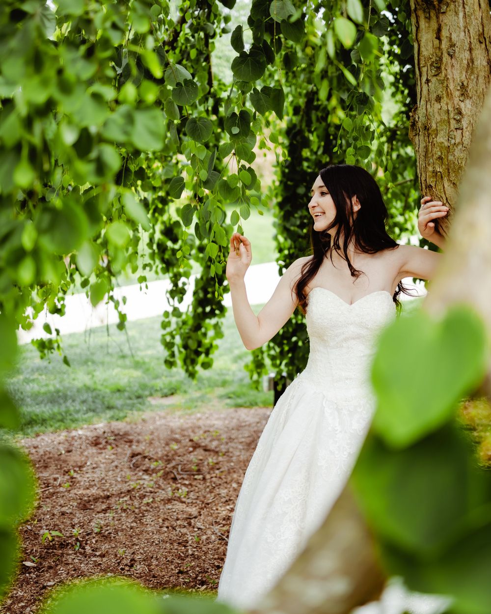 A white lace wedding dress photographed under drooping willow tree branches in a garden setting.