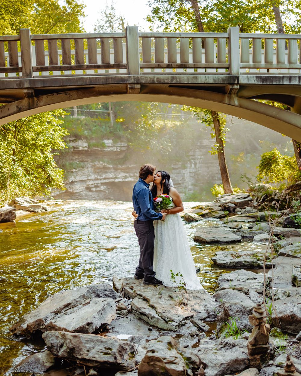A couple shares an intimate moment on rocky terrain beneath a wooden bridge over flowing water.