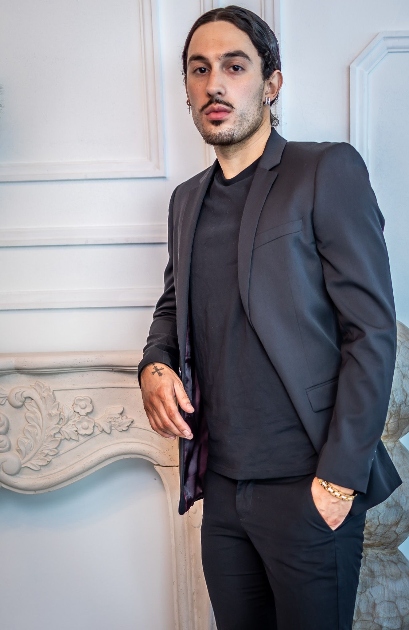 All-black formal suit ensemble photographed against white decorative wall.