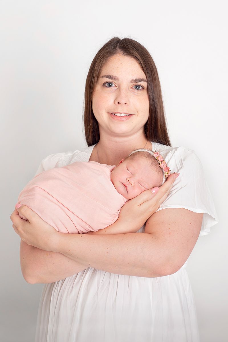 New mom in white dress proudly holds her newborn baby wrapped in a pink blanket