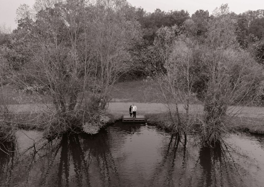 Black and white photograph of a solitary figure standing on a riverbank surrounded by bare trees.