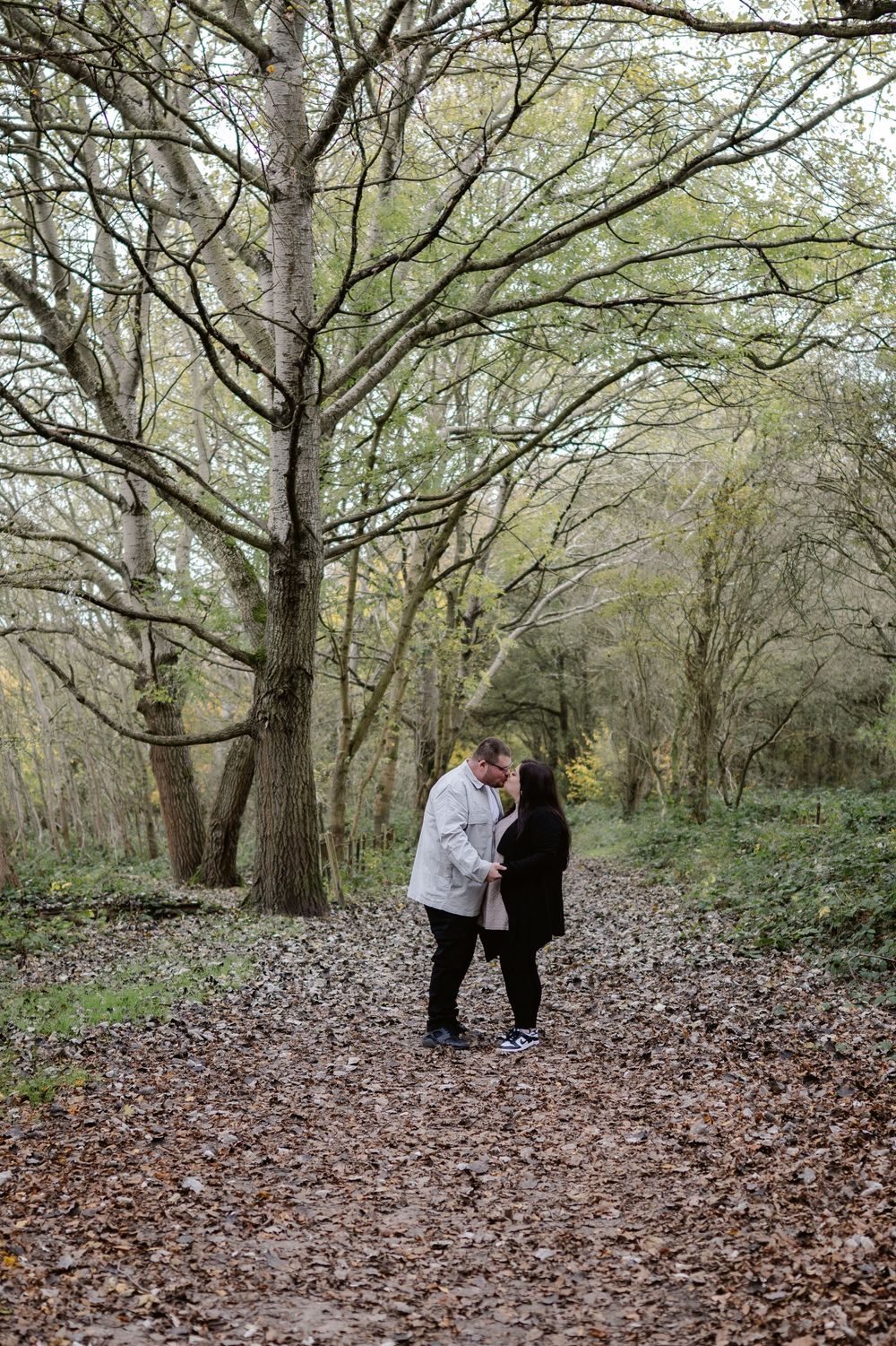 A couple standing on a leaf-covered forest path surrounded by tall bare trees in autumn.