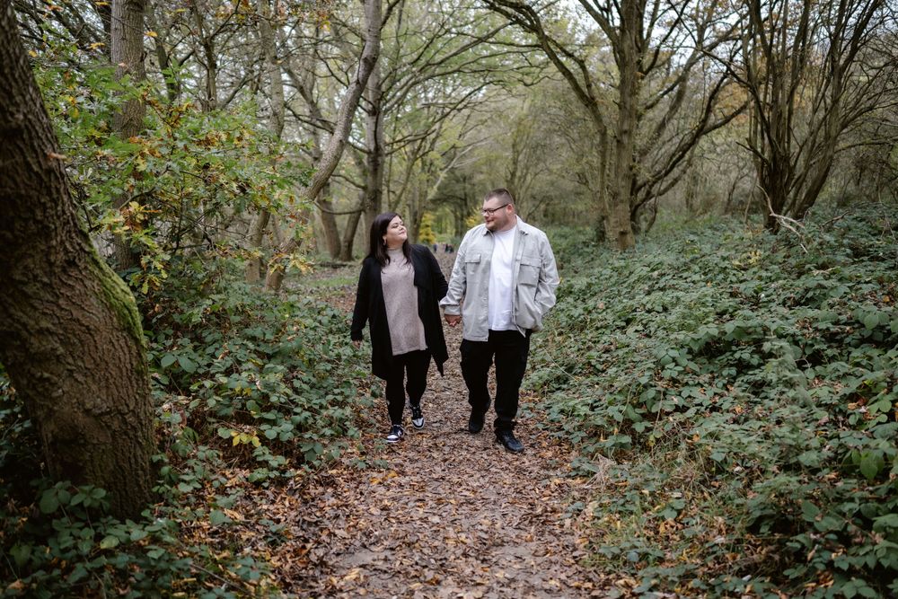 Two people walking together on a leaf-covered forest trail surrounded by green foliage and bare trees.