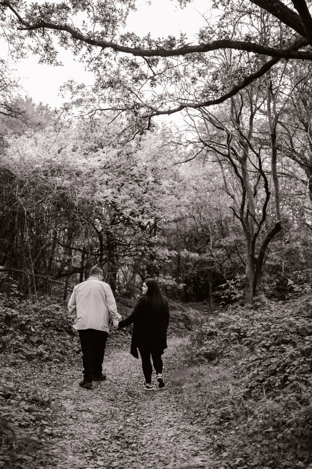 A black and white photograph of two people walking together on a wooded trail during autumn.