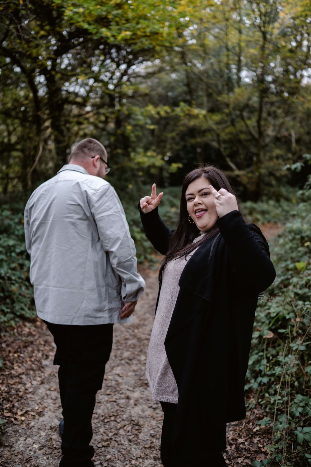 A playful couple poses on a wooded trail, with one making peace signs while the other faces away.
