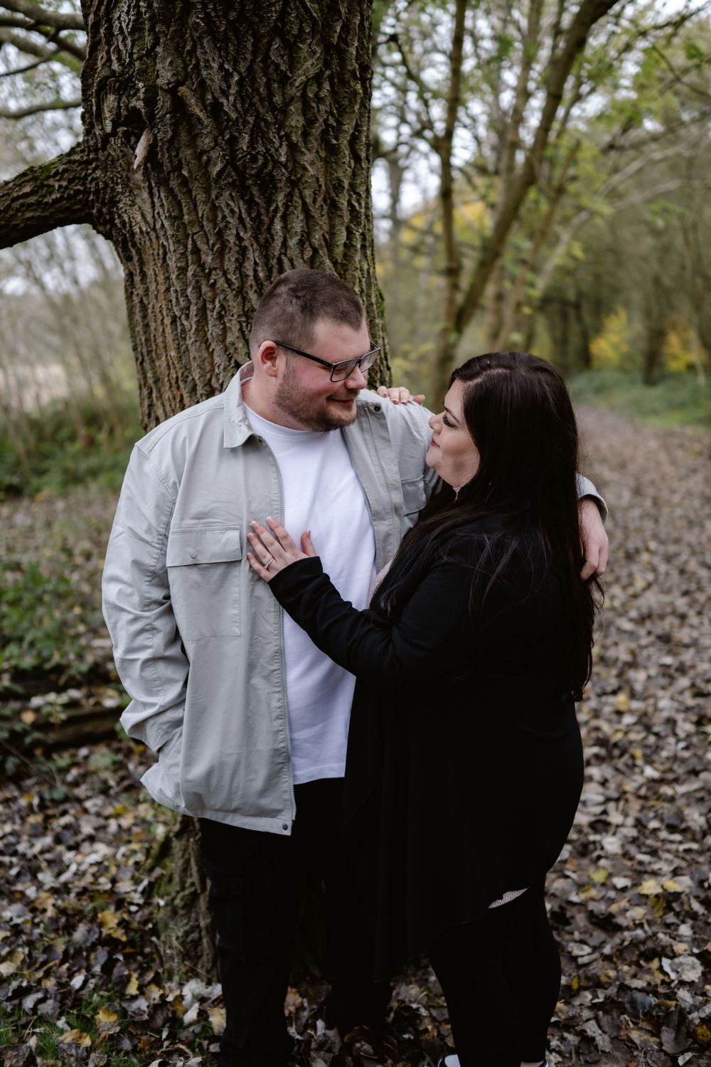 A couple shares an intimate moment while standing against a large tree trunk in a wooded area covered with fallen leaves.