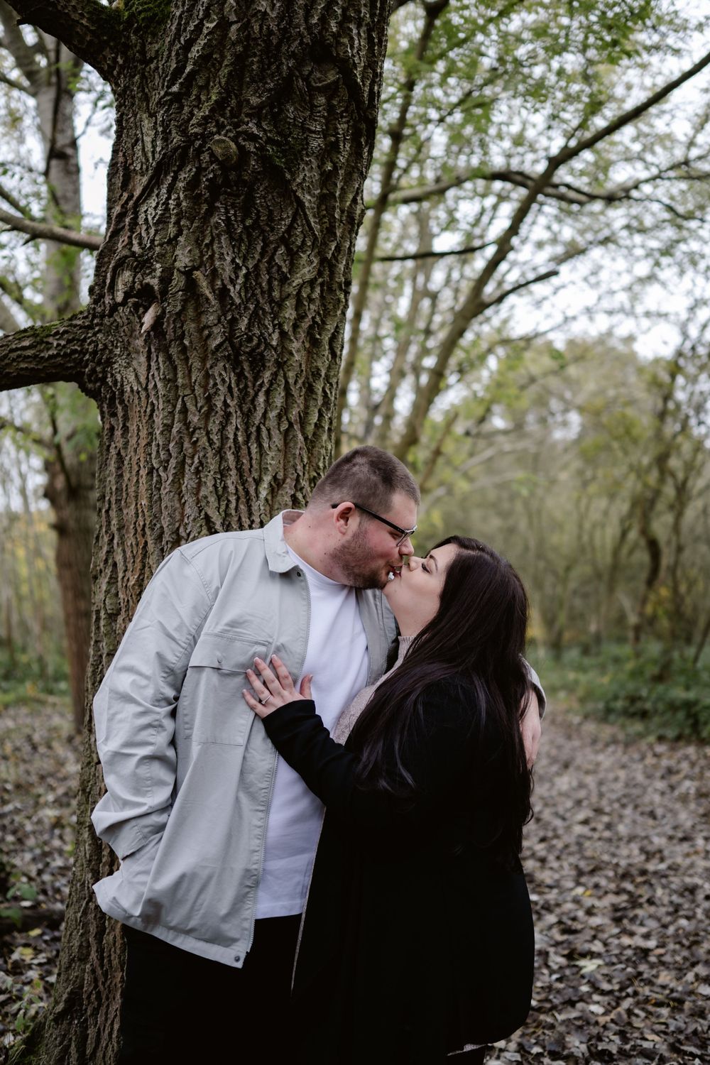 A romantic outdoor couple sharing a tender moment against a large tree trunk in a wooded setting.