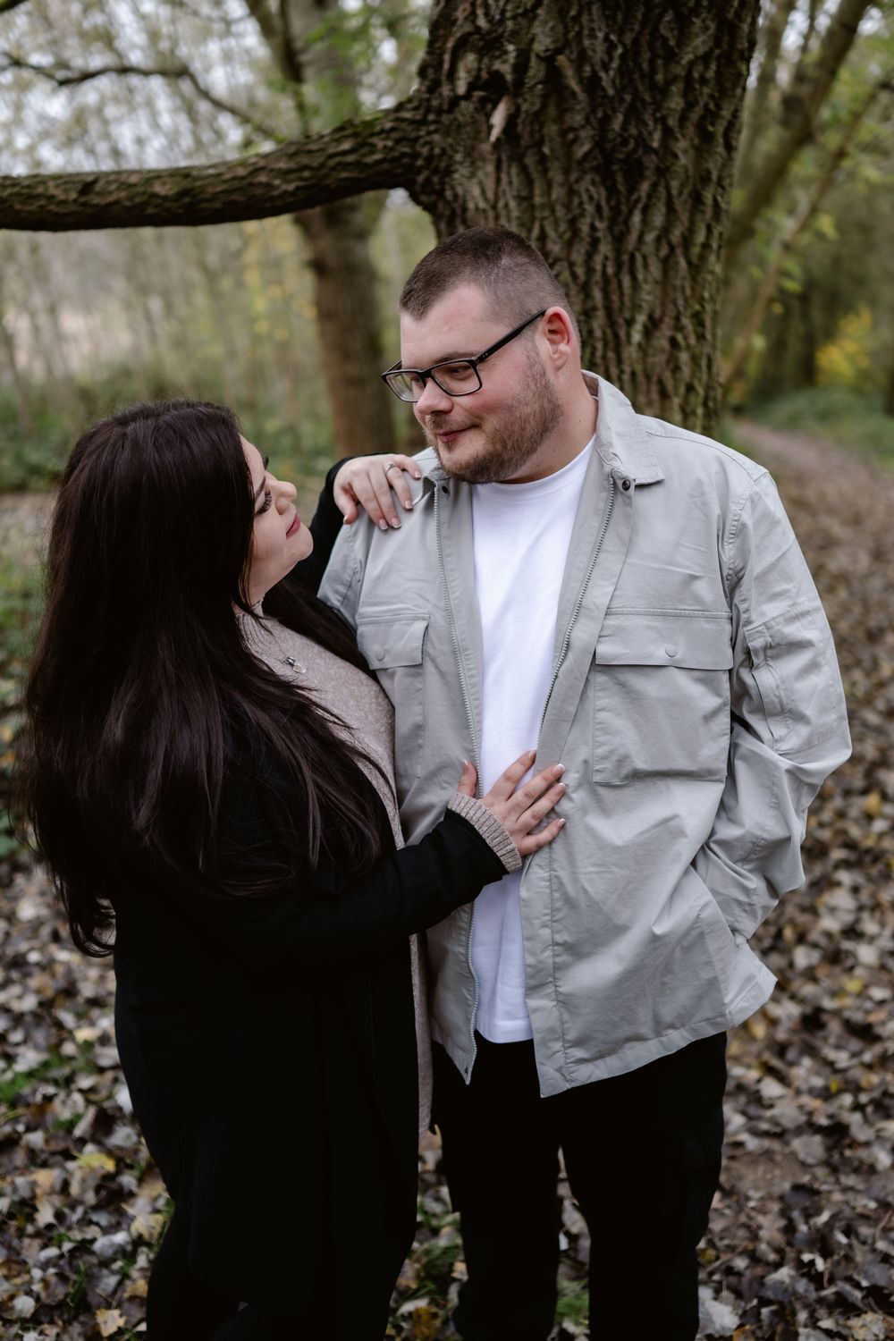 A couple shares an intimate moment while standing together beneath a large tree on an autumn day.