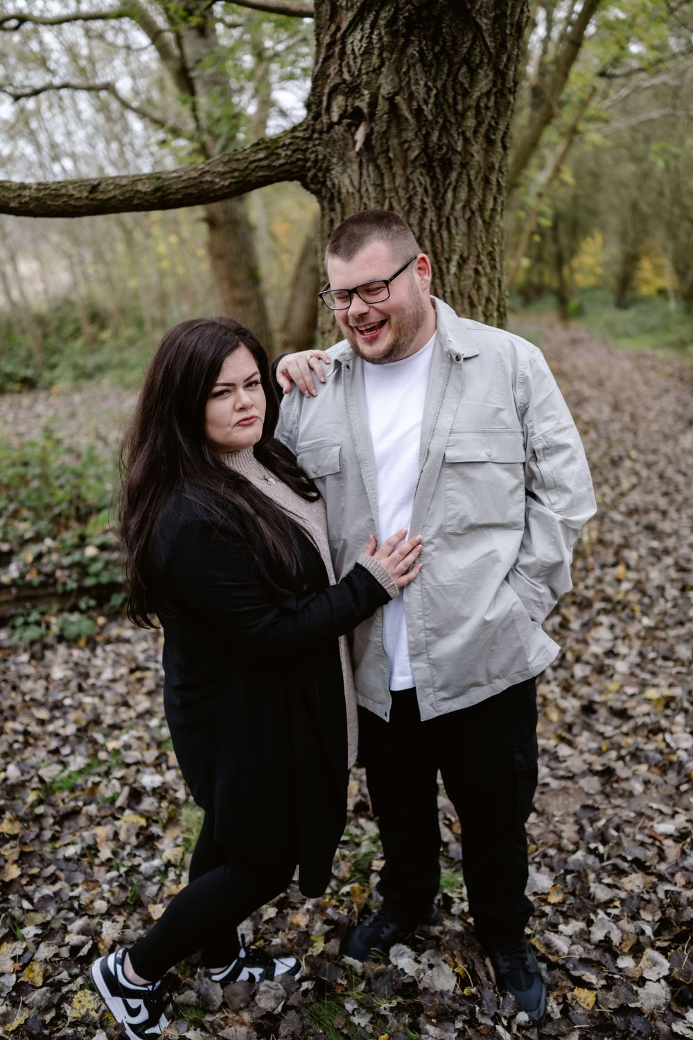 A loving couple embraces and smiles at each other while standing on autumn leaves in a forest setting.