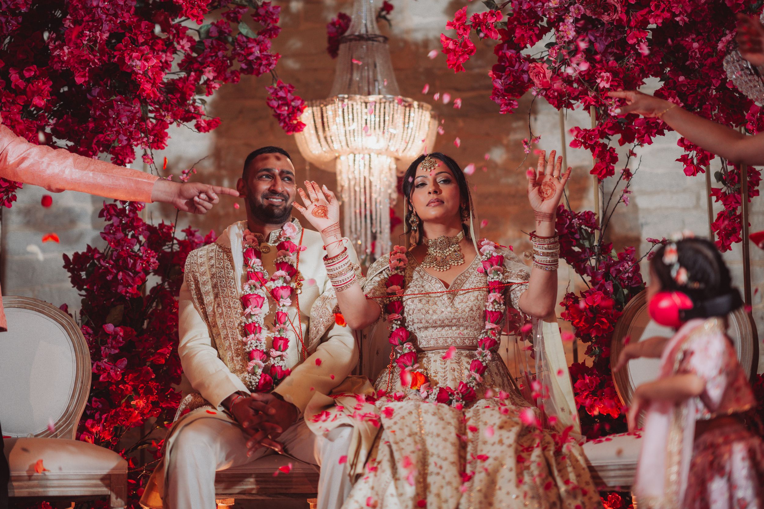 Elegant Indian wedding ceremony setup with red floral decor and crystal chandelier featuring couple in traditional attire.