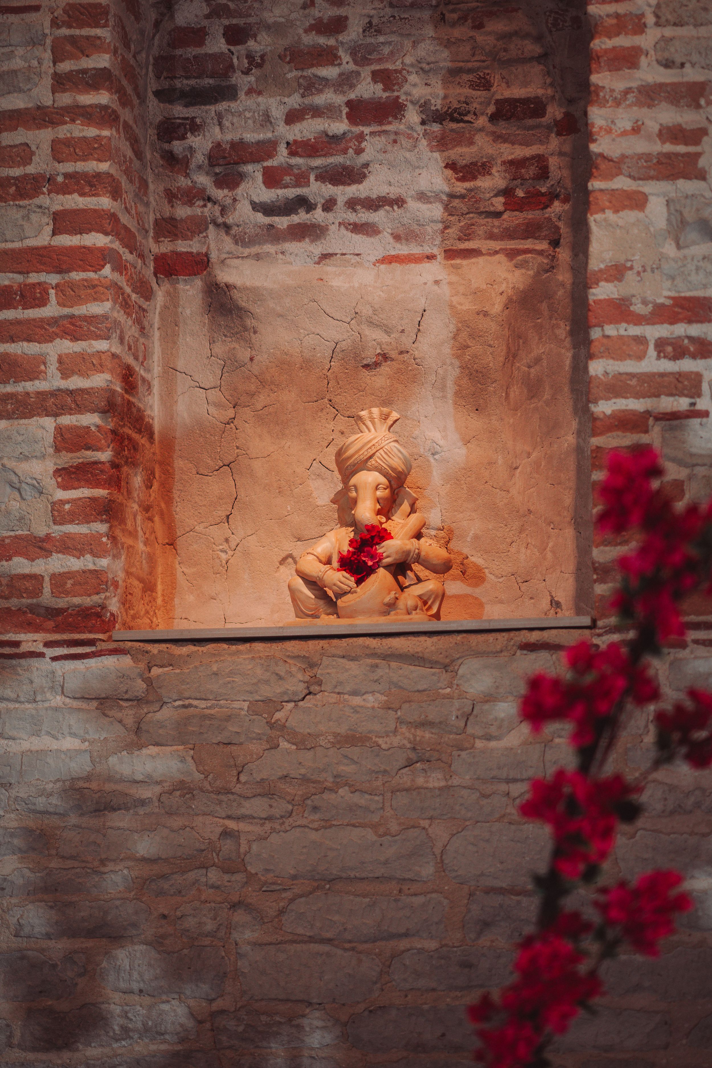 Clay cherub sculpture in brick alcove with red bougainvillea flowers in foreground.
